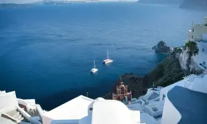 Vue d'un village blanc sur une falaise surplombant la mer Égée. Deux voiliers naviguent paisiblement.