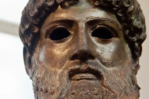 Tête en bronze de Zeus, dieu de la foudre, avec une barbe et des cheveux bouclés. Son regard est intense, prêt à lancer ses éclairs.