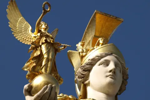 Statue dorée d'une déesse de la guerre ailée, tenant une couronne et une lance, perchée sur une sphère. Une autre tête de déesse ornée d'un casque doré est visible.