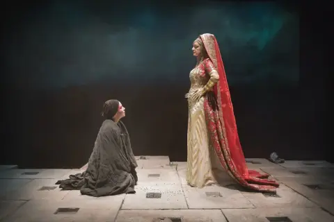 Sur scène, une femme agenouillée, drapée de noir, implore une figure royale vêtue d'or et de rouge. Une scène du mythe d'Électre.