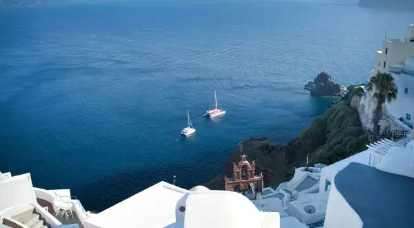 Vue d'un village blanc sur une falaise surplombant la mer Égée. Deux voiliers naviguent paisiblement.