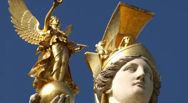 Statue dorée d'une déesse de la guerre ailée, tenant une couronne et une lance, perchée sur une sphère. Une autre tête de déesse ornée d'un casque doré est visible.