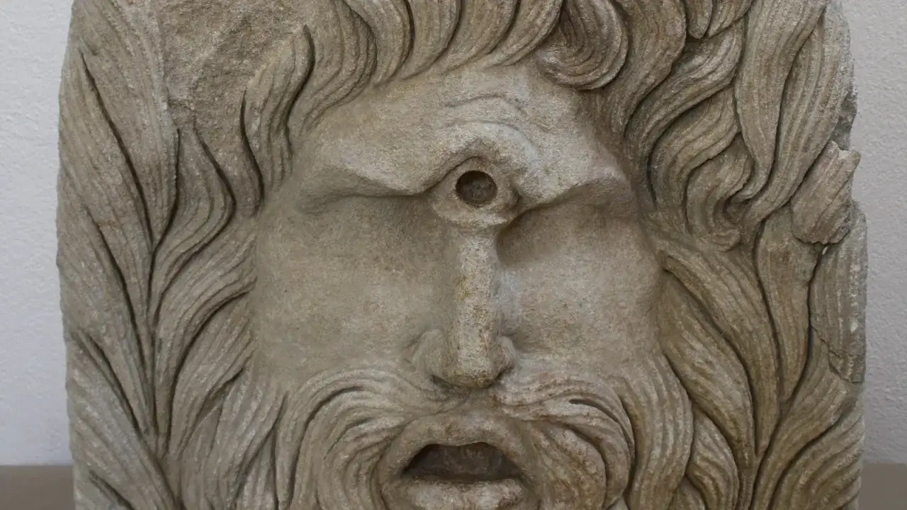 Masque de pierre d'un cyclope, avec une chevelure ondul&eacute;e et une barbe fournie. Son unique &oelig;il est creus&eacute; au centre du front.