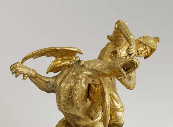 Statue dorée d'un guerrier combattant un dragon, évoquant le roi mythique d'Iolcos. Le guerrier, casqué, saisit la gueule ouverte de la créature ailée.
