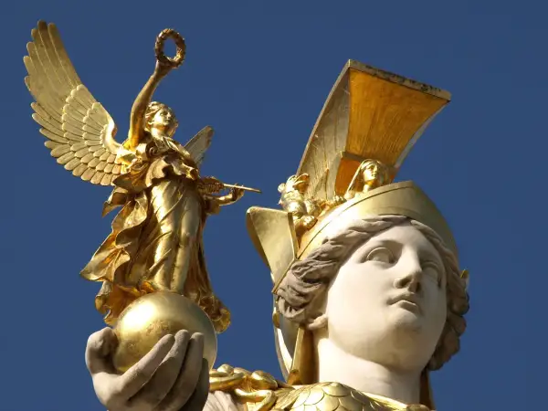 Statue dorée d'une déesse de la guerre ailée, tenant une couronne et une lance, perchée sur une sphère. Une autre tête de déesse ornée d'un casque doré est visible.
